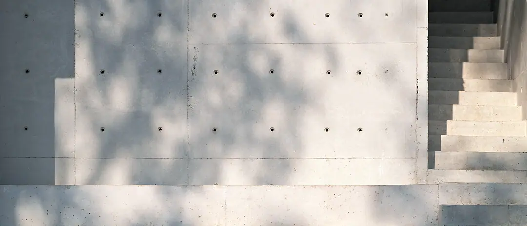 A smooth concrete wall with tie holes beside a concrete stair, dappled leaf shadows across it